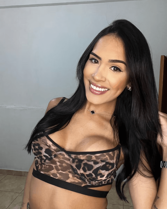 Portrait of a smiling woman with long black hair, wearing a leopard-print top and necklace, posing for a selfie indoors.