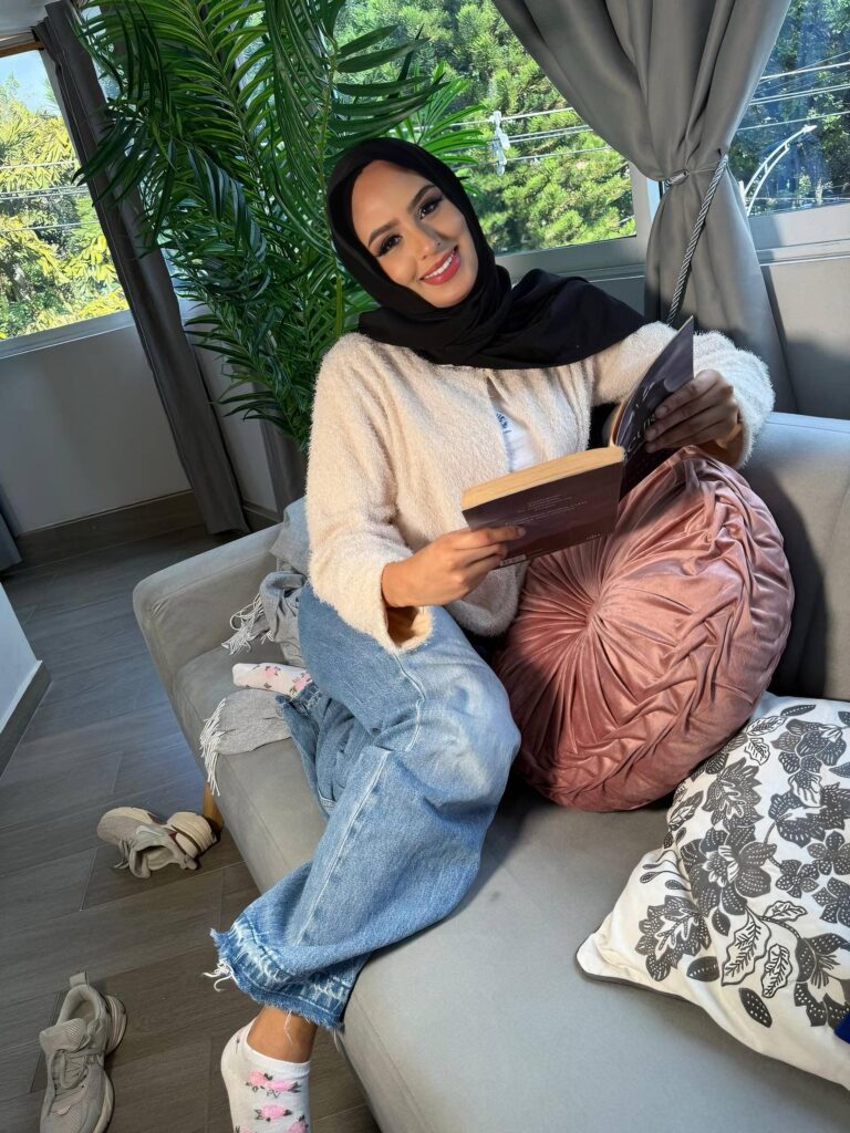 Woman in a black hijab and beige cardigan sits on a gray sofa, reading a book with a smile, indoors by a window with plants in the background.