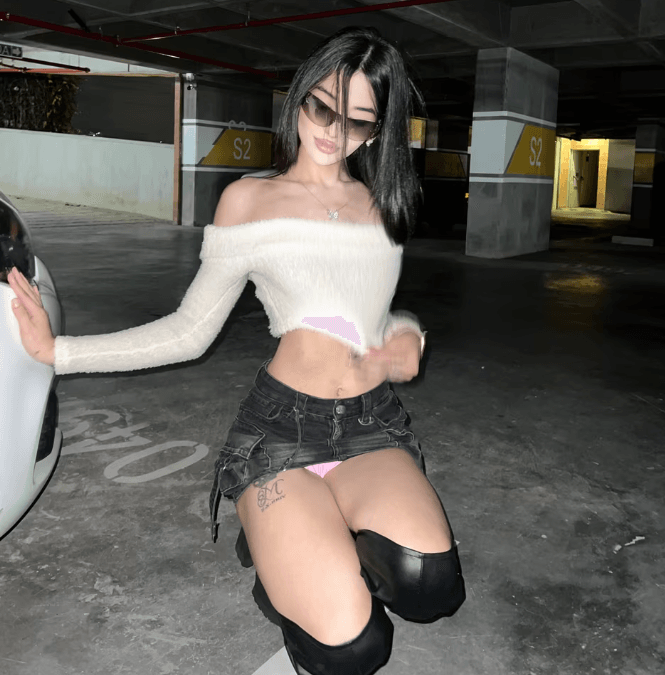 Young woman in an off‑the‑shoulder white sweater and black denim shorts kneels beside a car in a parking garage; pink underwear is visible.