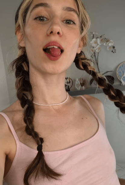 Young woman with two braided pigtails, wearing a pearl choker and pink tank top, sticking out her tongue playfully.