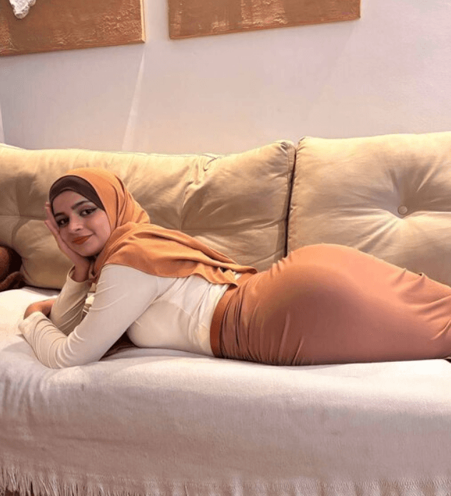 Woman wearing an orange hijab and white top lying on a beige sofa, propped on her elbow and smiling at the camera.