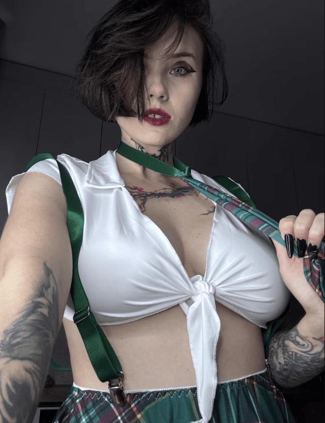 Selfie of a woman with short dark hair, bold makeup, wearing a white knotted crop top with green suspenders and a plaid skirt, showing tattoos on her chest and arms