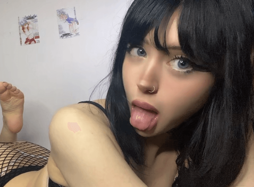 Person with dark hair and nose ring sticking out tongue at the camera, wearing a black top and fishnet stockings.
