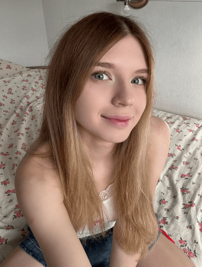 Young woman with long light brown hair and blue eyes sitting on a floral bedspread, smiling at the camera.