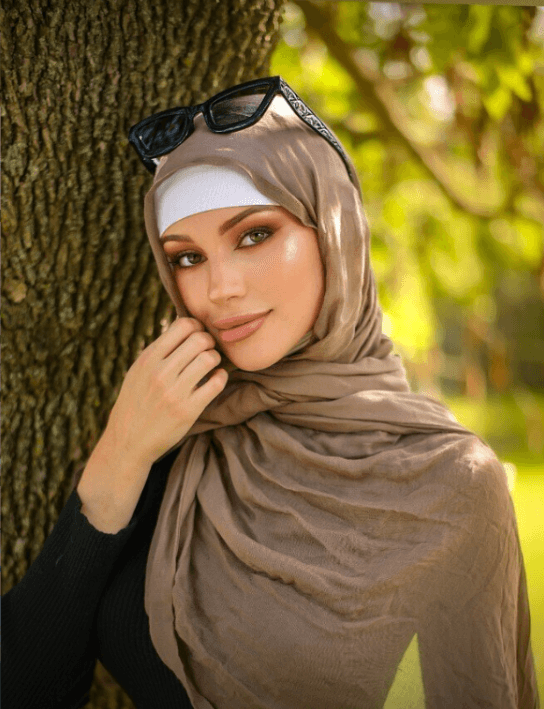 Woman wearing beige hijab and white headband, sunglasses perched on her head, smiling beside a tree in a sunny park.