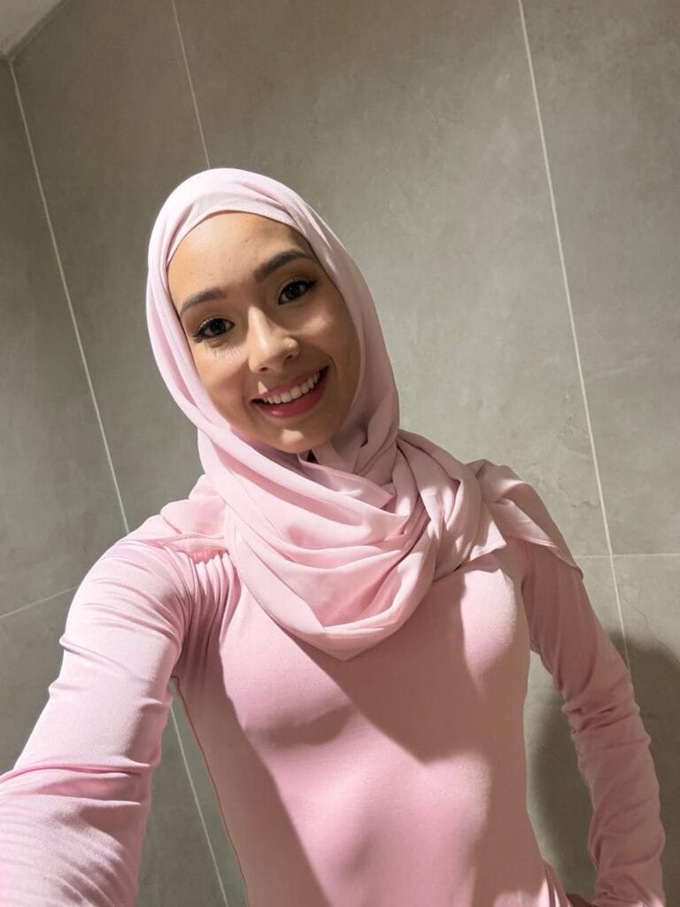 Smiling woman in a light pink hijab and matching top taking a selfie in a tiled indoor wall setting.
