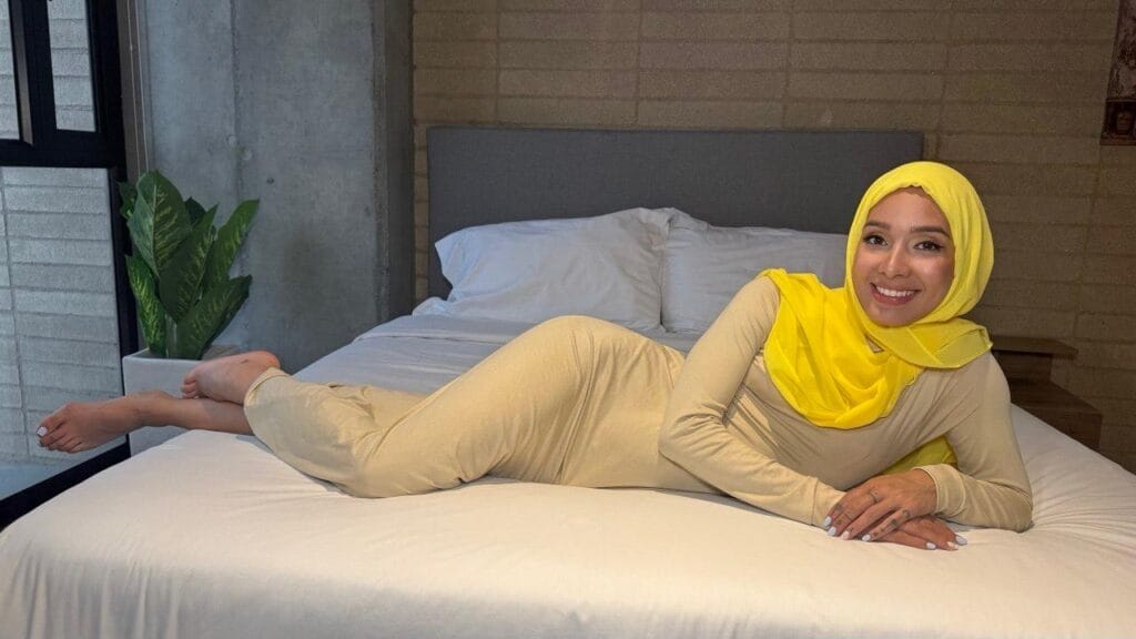 Woman in a beige outfit and bright yellow hijab lounging on a bed, smiling at the camera in a modern bedroom.