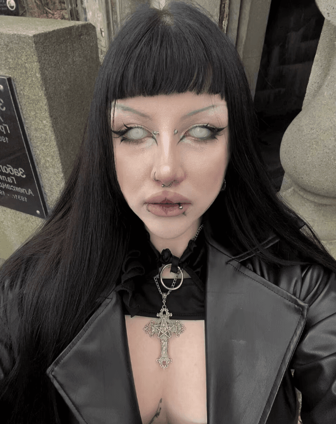 Person with long black hair and blunt bangs wearing a black leather jacket, gothic makeup, multiple facial piercings, and a large ornate cross necklace.