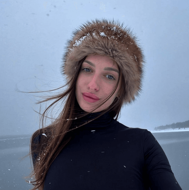 Woman in a black turtleneck with fur-lined hood outdoors in snowfall, facing the camera.