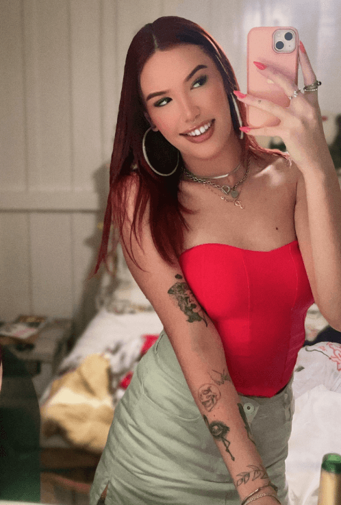 Woman taking a mirror selfie with a pink phone, wearing a red strapless top and light green skirt, tattoos on her arm and hoop earrings.