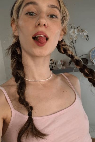 Young woman with two braided pigtails wearing a pink tank top and a pearl necklace, sticking her tongue out playfully at the camera.