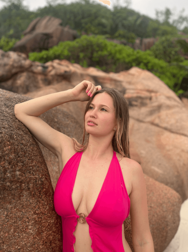 Woman in a pink halter swimsuit leaning against rocky beach formations, looking up thoughtfully.