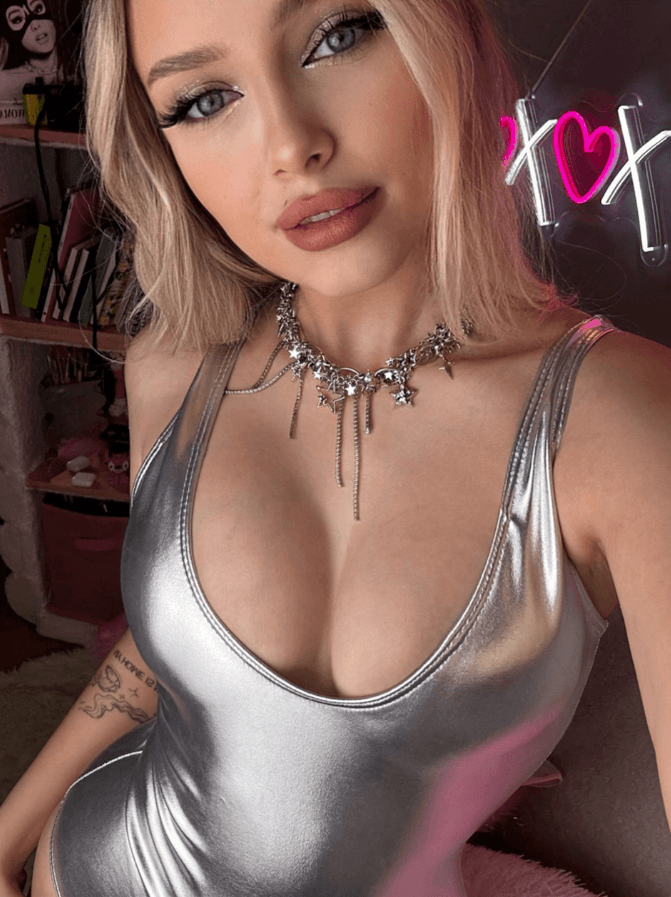 Selfie of a woman in a shiny silver bodysuit wearing a starry necklace; pink neon sign and tattoo visible in the background.