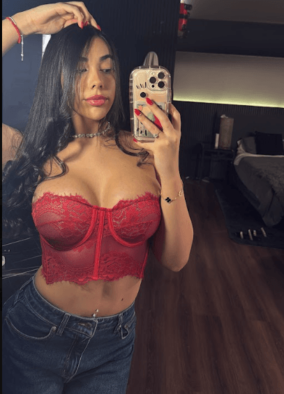 Woman taking a mirror selfie in a red lace bustier and blue jeans, hair down, in a dimly lit bedroom bathroom area.