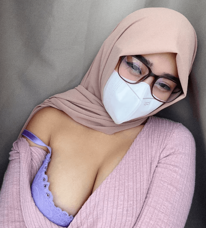 Person in a beige hijab, glasses, and a white mask, taking a selfie in a pink knit top with lavender lace bra showing.