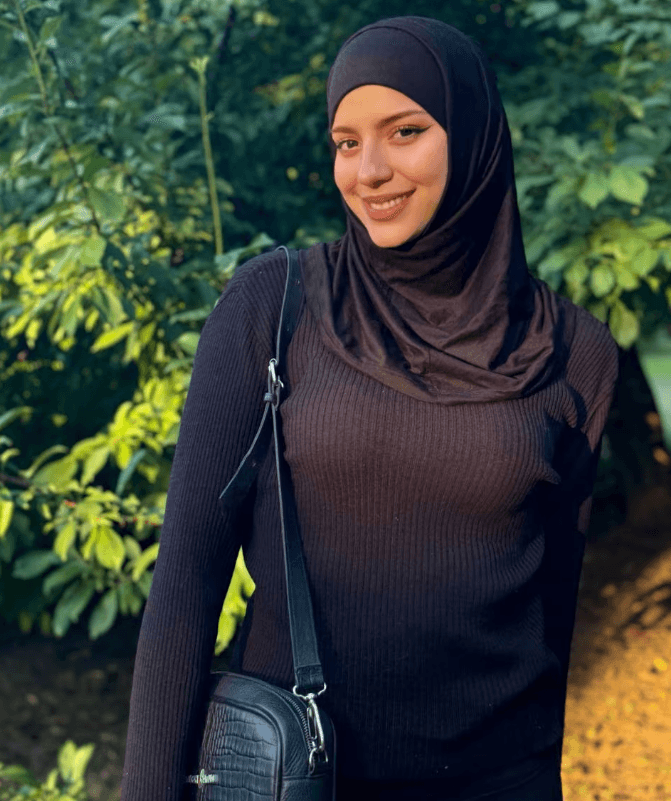 Smiling woman in a black hijab and ribbed sweater outdoors, with a crossbody bag, in a green, sunlit garden/poster-friendly setting.