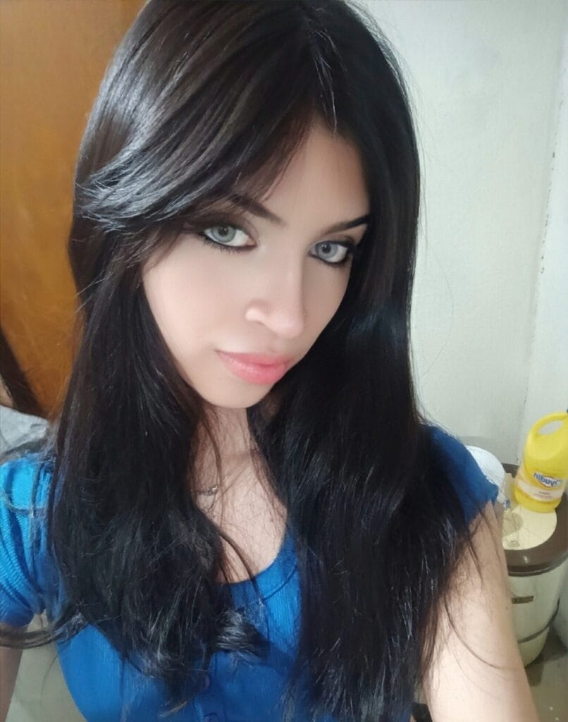 Close-up of a woman with long dark hair, blue top, looking toward the camera with a neutral expression in an indoor setting.