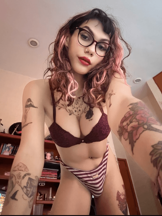Selfie of a person with pink-tinted hair and glasses wearing a burgundy bra and striped panties, showing tattoos on arms and torso in a cluttered room.