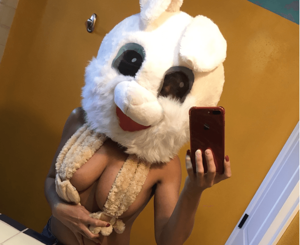 Person in a large white bunny head mask, topless with a furry scarf, taking a mirror selfie with a red phone against a yellow wall.