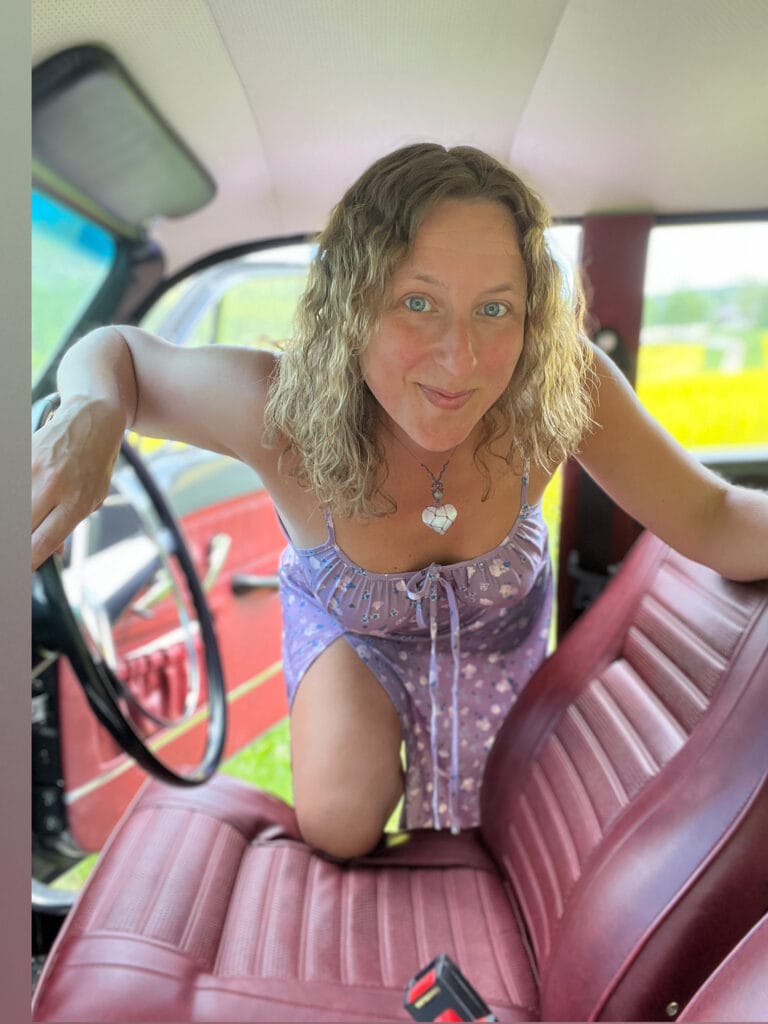 Woman leaning toward camera inside a red car, wearing a purple floral dress and a heart-shaped necklace.