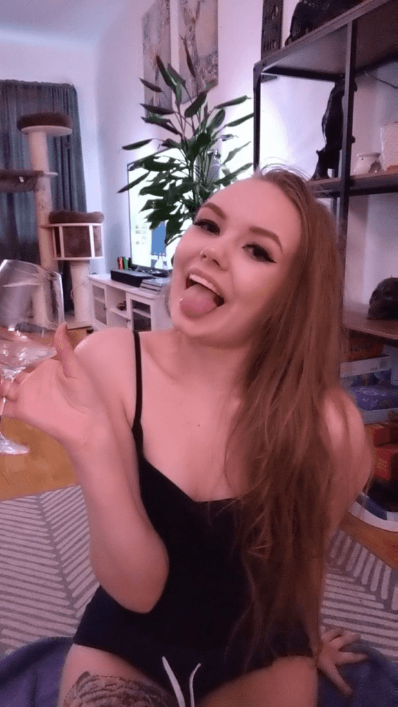 Young woman in a black tank top playfully sticking out her tongue while holding a wine glass, indoors in a cozy room with plants and shelves.