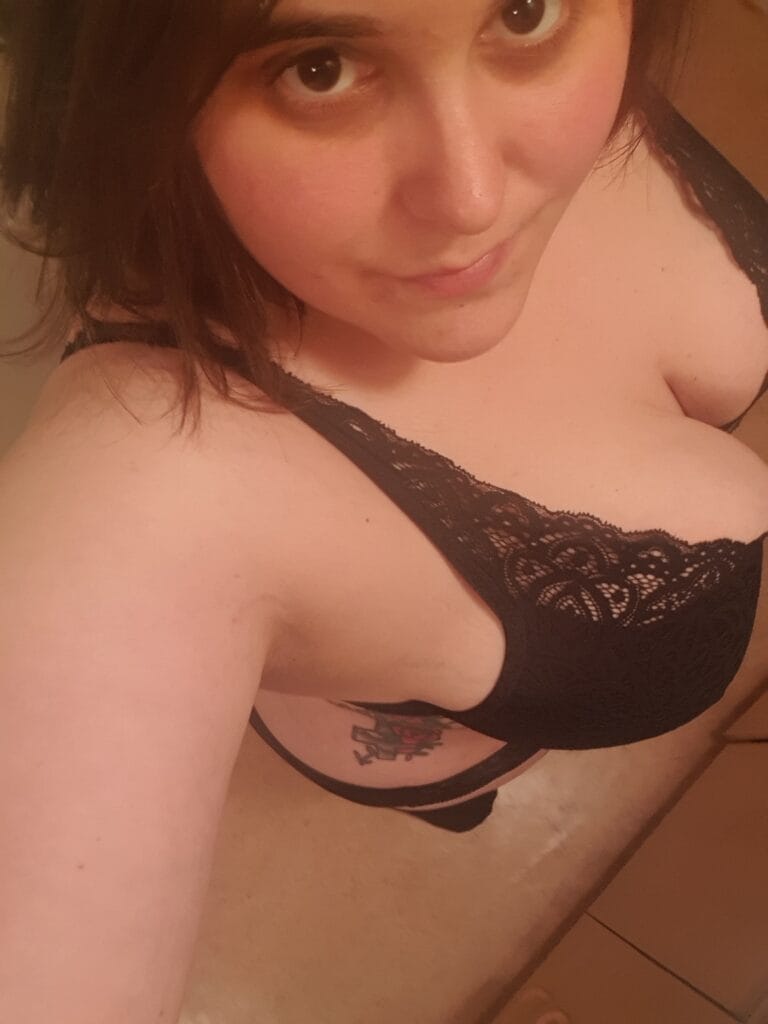 Close-up of a person wearing a black lace bra, looking up at the camera, with a tattoo visible on the side rib area.