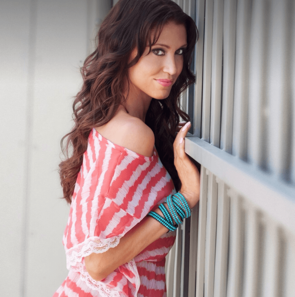 Portrait of a woman with long brown hair wearing a pink and white off-shoulder dress, leaning against a railing with turquoise bracelets on one wrist.